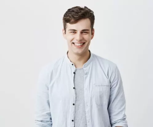 Handsome attractive young man dressed in light blue shirt over t-shirt with dark hair and appealing blue eyes looknig at camera, smiling broadly, demonstrating white teeth, being happy and pleased. Handsome attractive young man dressed in light blue shirt over t-shirt with dark hair and appealing blue eyes looknig at camera, smiling broadly, demonstrating white teeth, being happy and pleased, posing indoors. Positive emotions